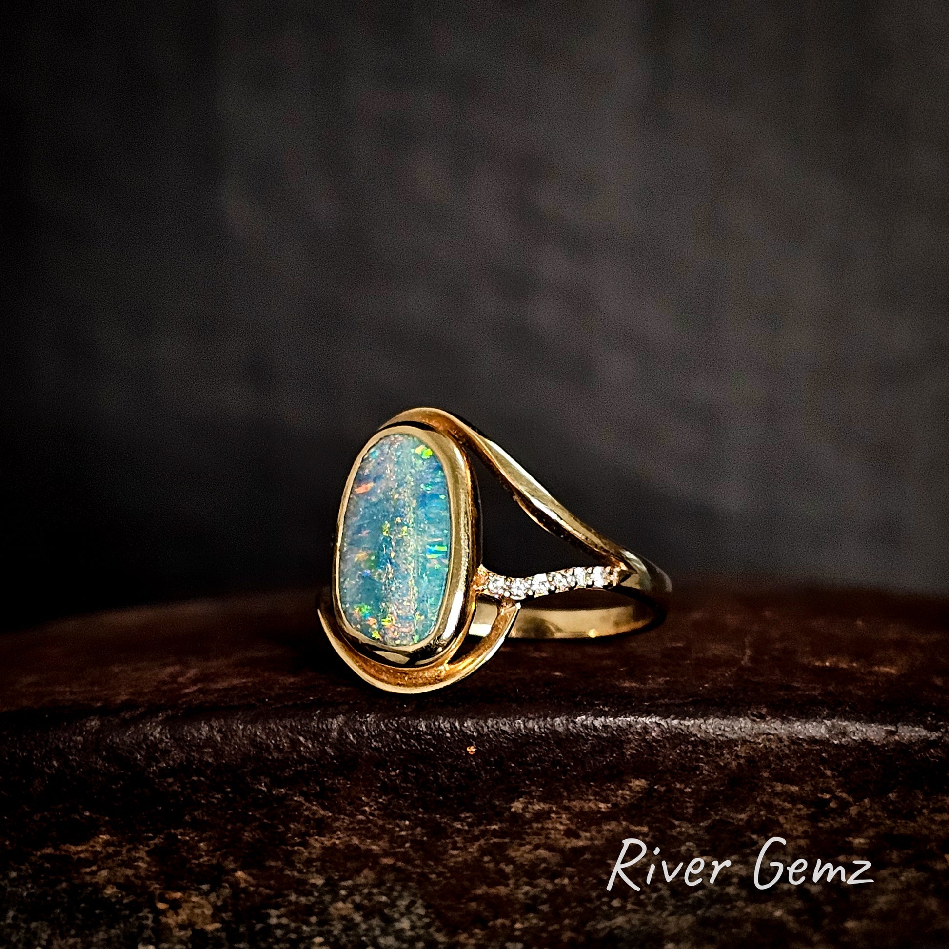Gold ring with a multi-coloured opal stone and diamonds on a dark background, featuring the brand 'River Gemz'.