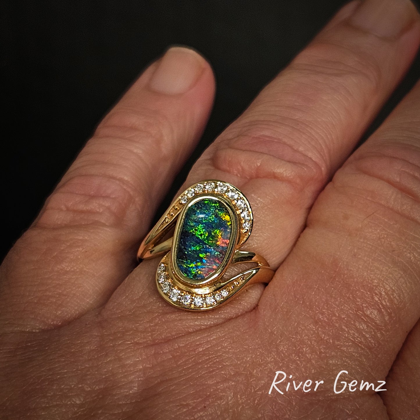 Gold ring with an opal stone and multiple diamonds on a hand, branded 'River Gemz'.