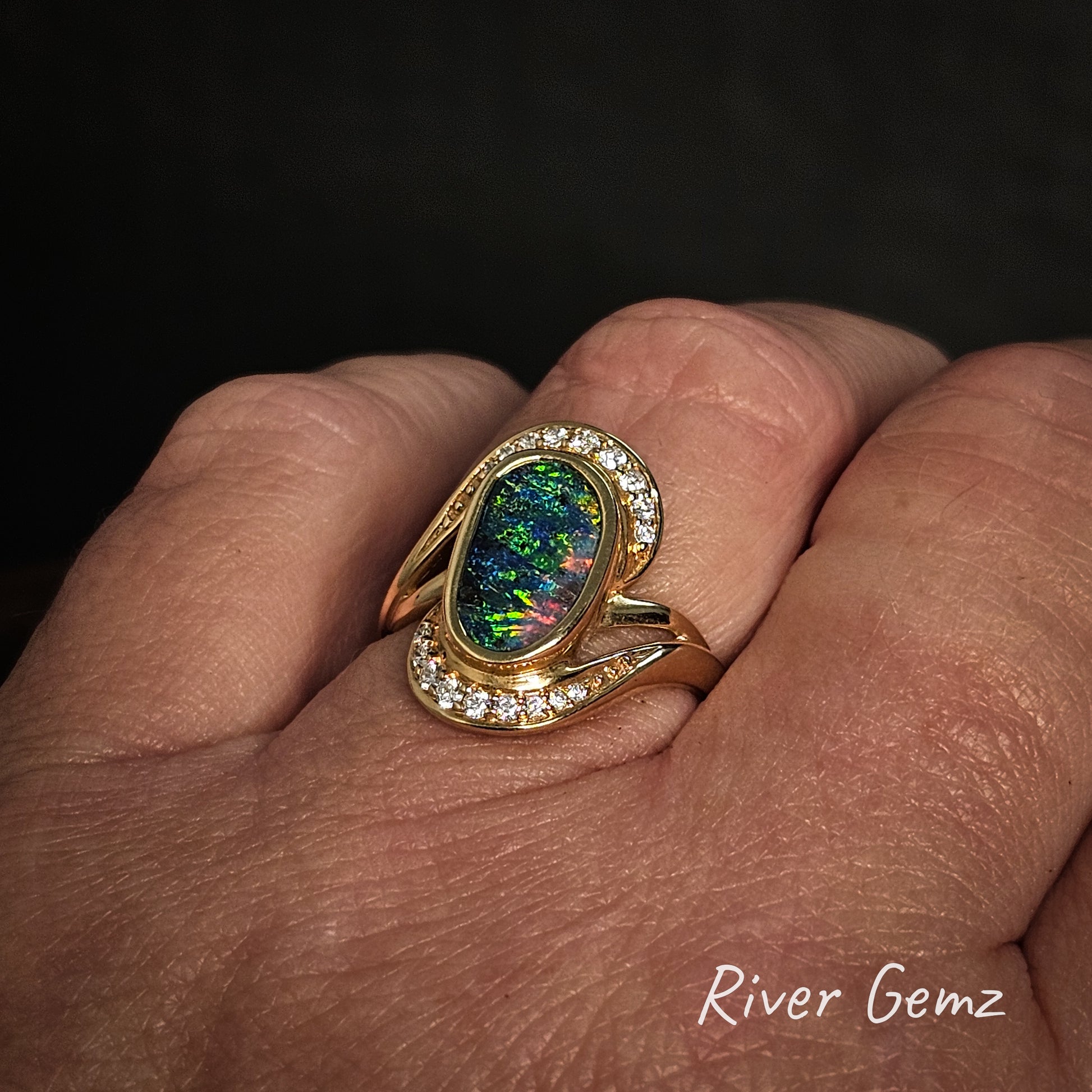 Gold ring with a colorful gemstone on a hand, branded 'River Gemz'.