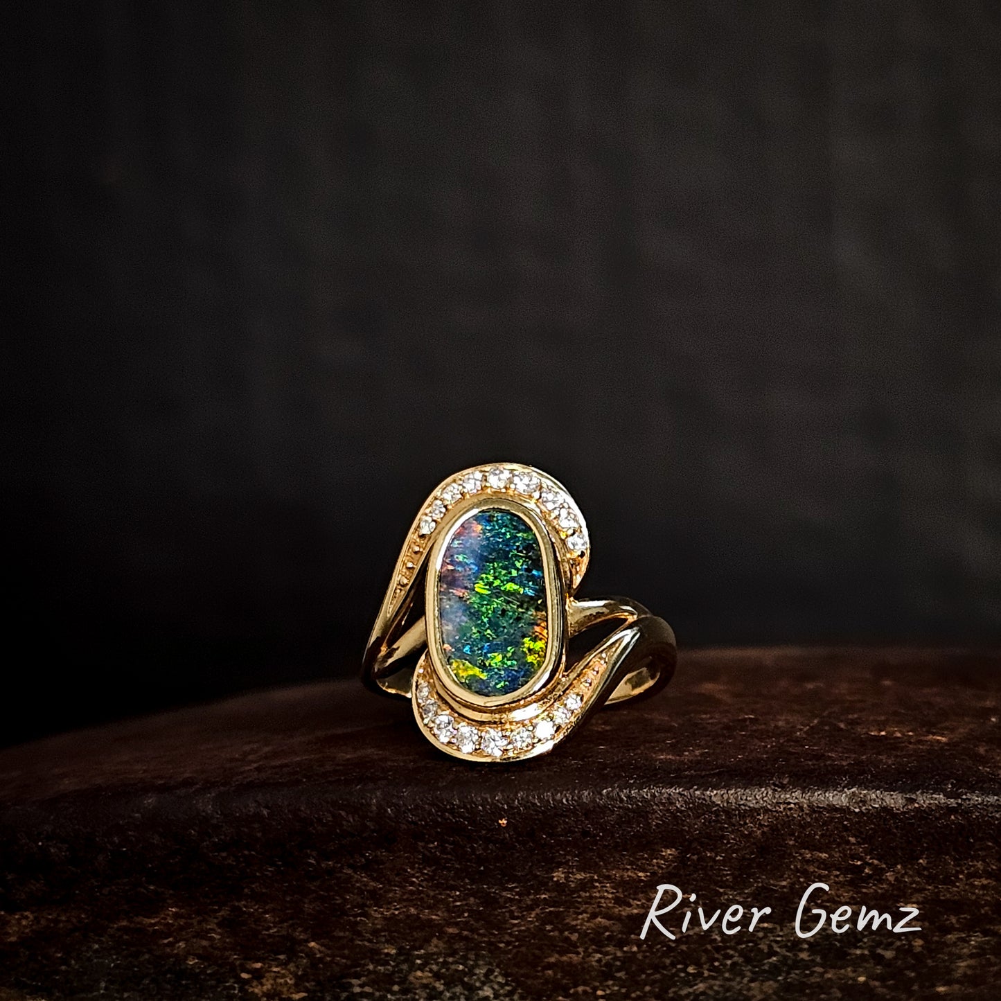 Gold ring with a multi-coloured boulder opal and diamonds on a dark background, featuring the brand 'River Gemz'.