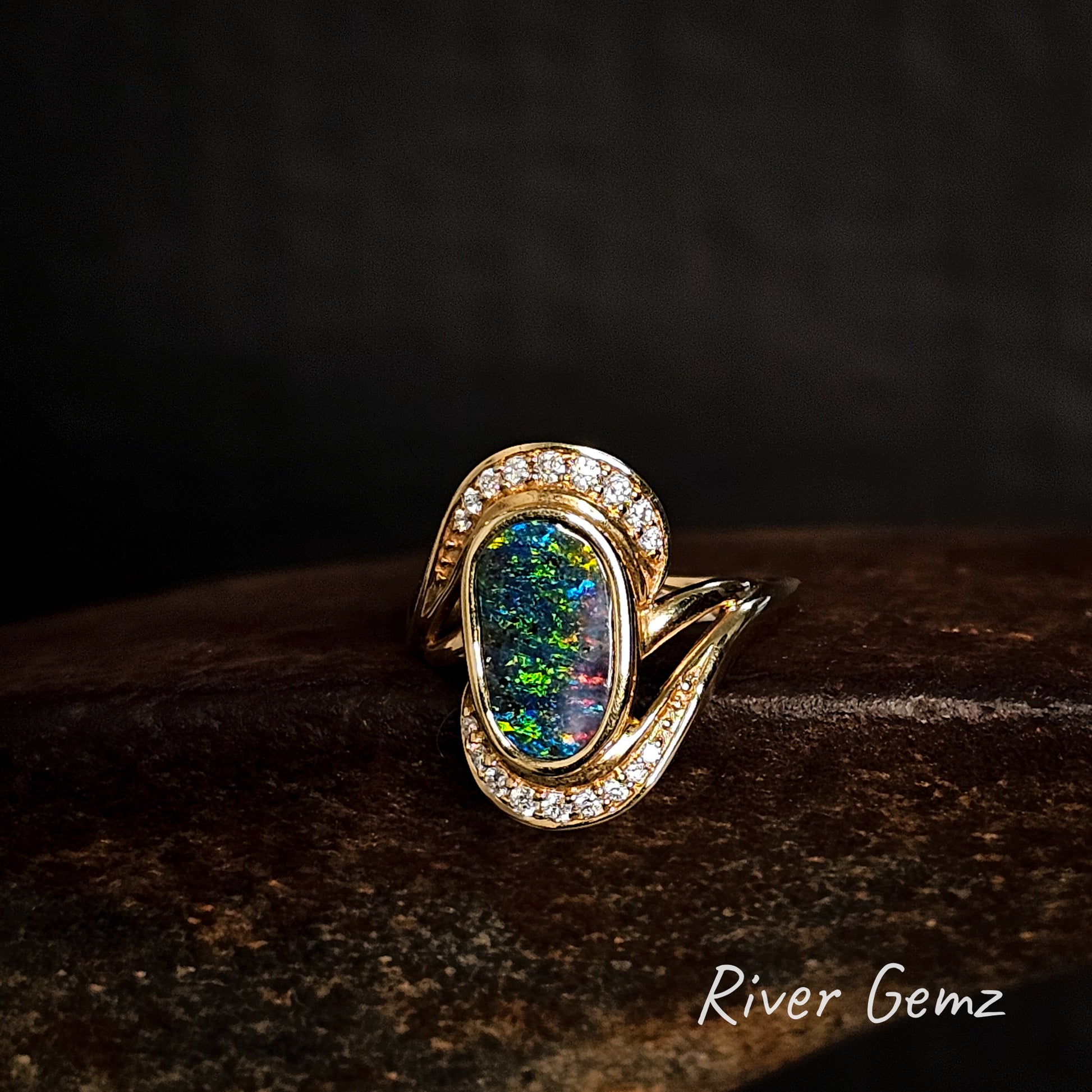 Gold ring with a colorful gemstone and channel set diamonds above and below the opal on a dark background, branded 'River Gemz'.
