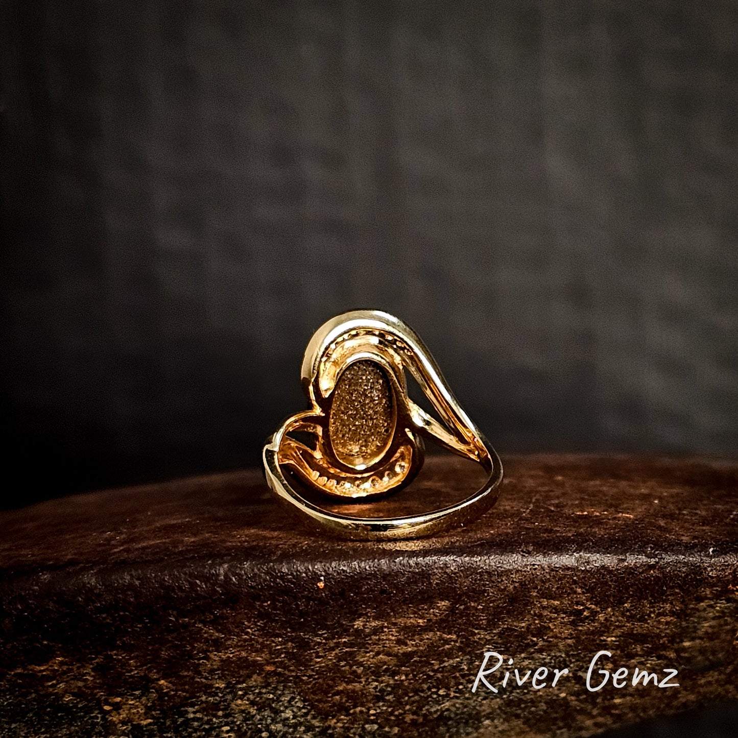 Back of gold ring showing the ironstone matrix of the included boulder opal on a cast iron surface with a dark background, featuring the brand 'River Gemz'.
