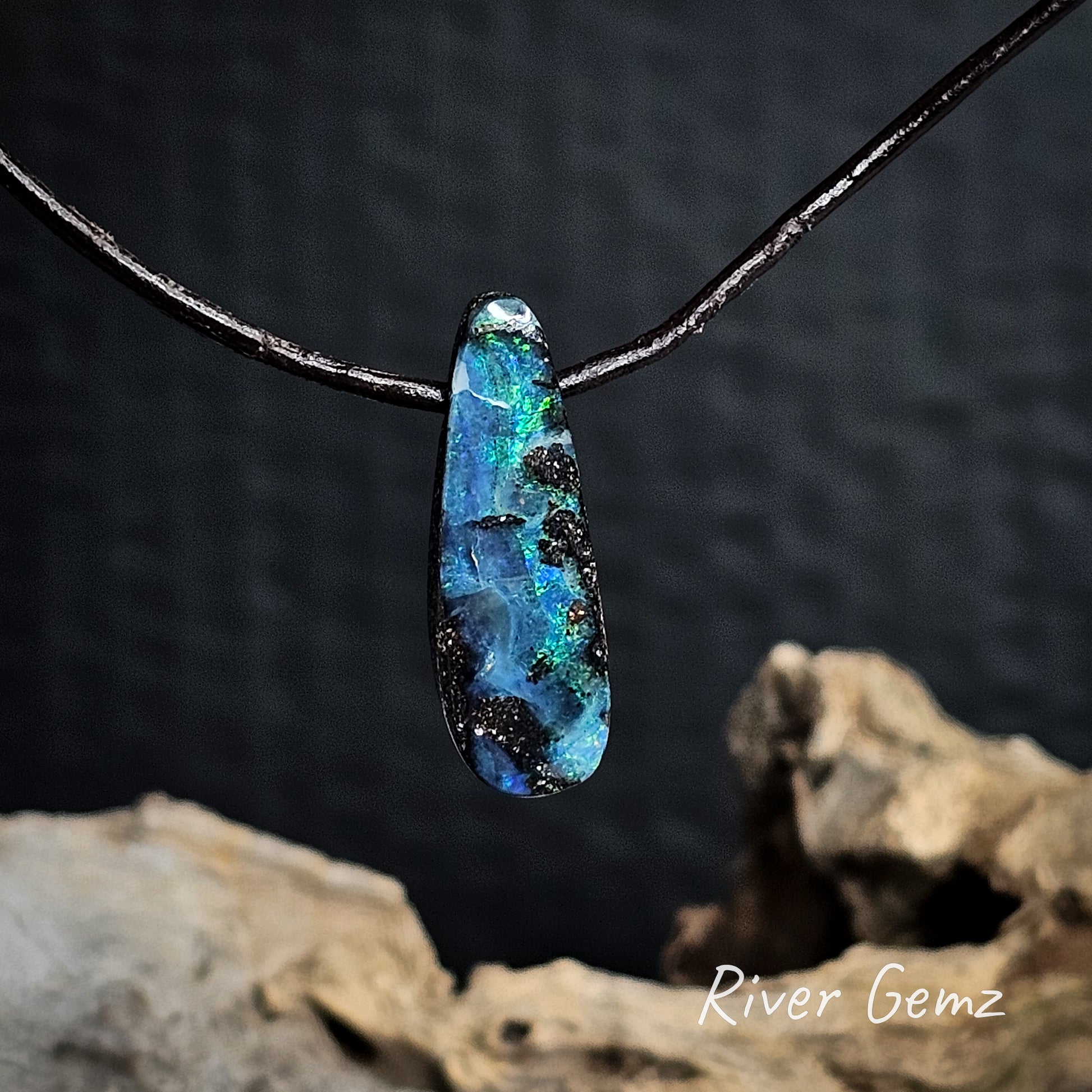 Blue and green opal on a dark brown cord with a blurred natural background.