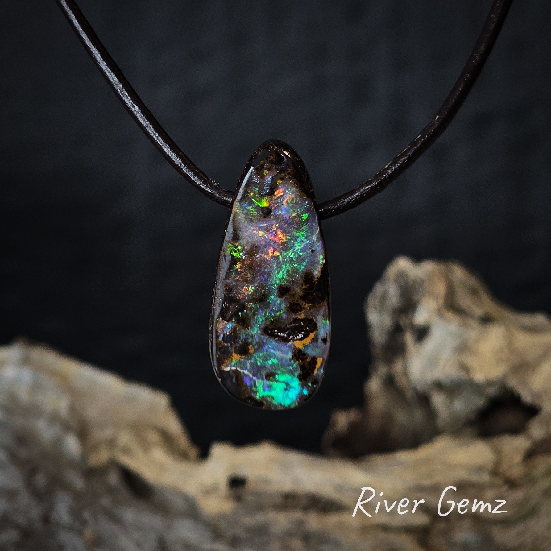 Opal pendant on a dark cord with a blurred natural wooden background, featuring the brand 'River Gemz'.