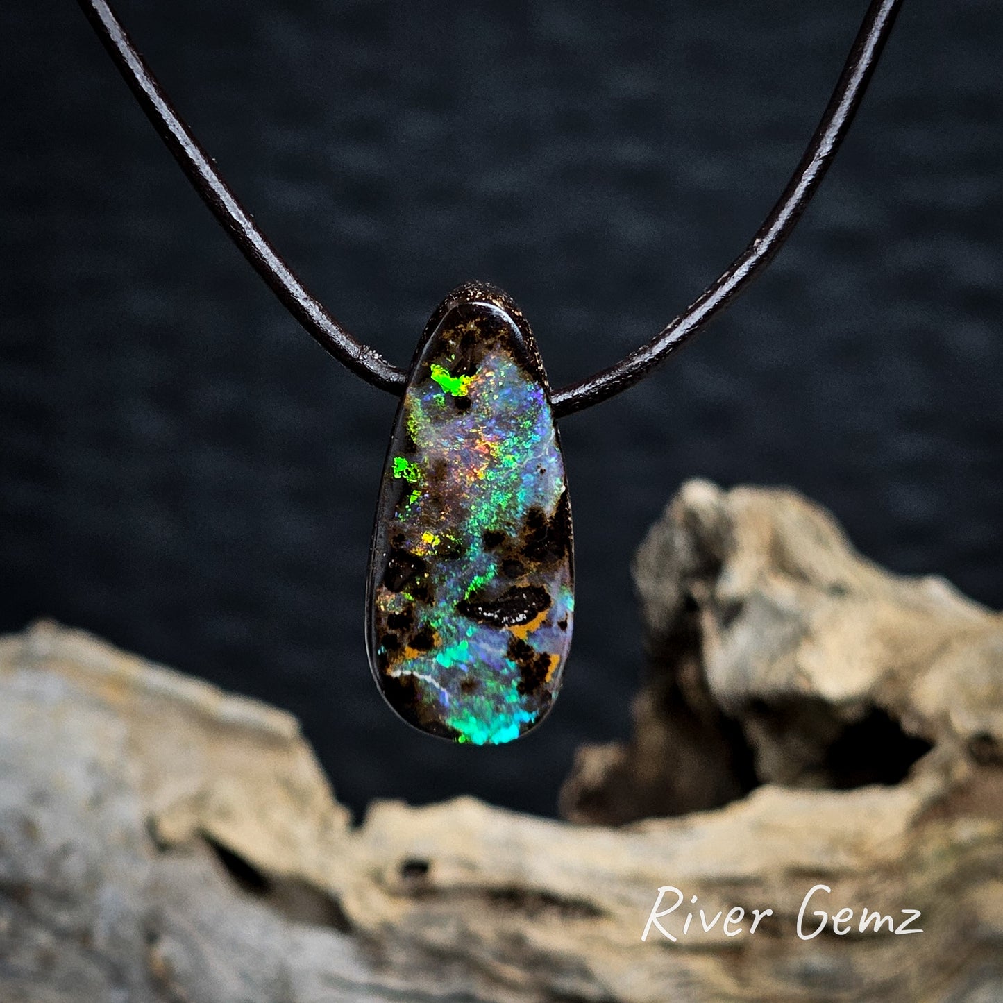 Tear-drop shaped opal pendant on a brown cord with a dark background and 'River Gemz' branding.