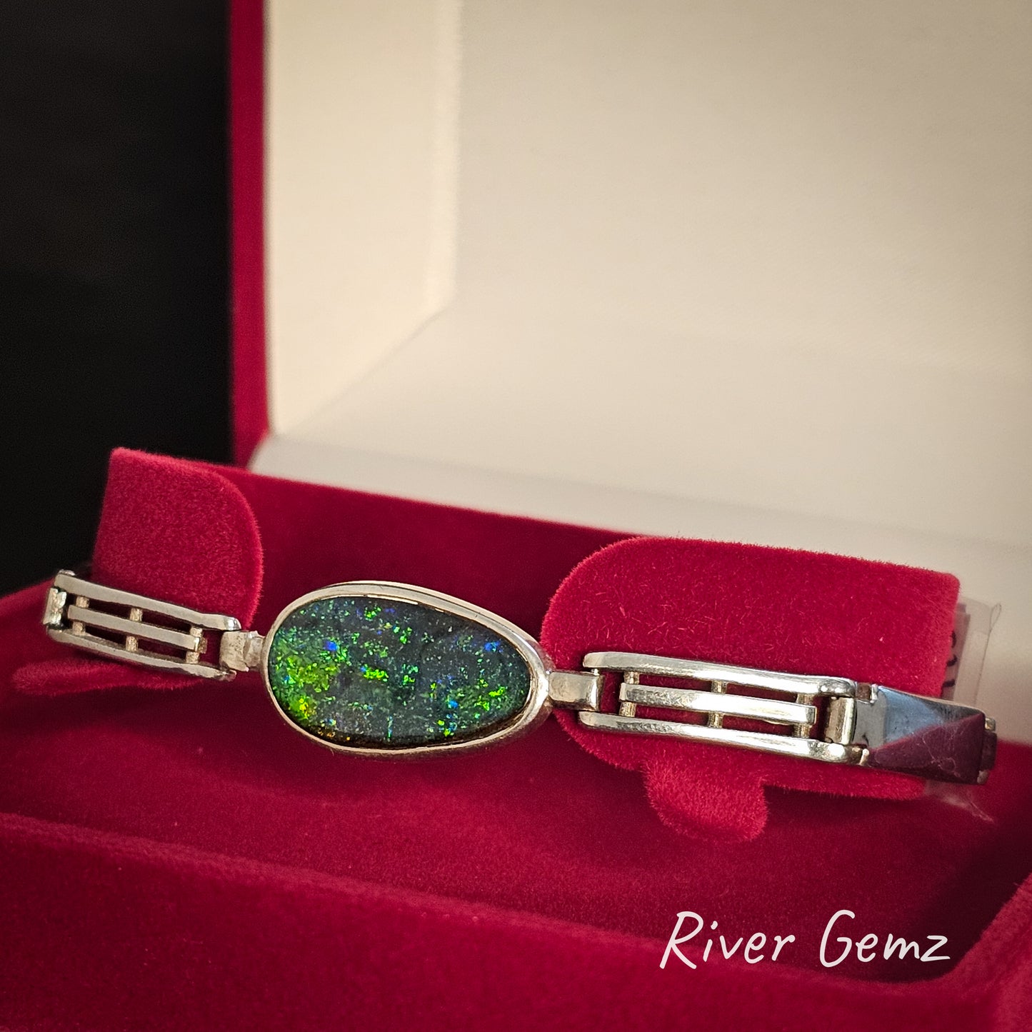Silver bracelet with a colorful gemstone on a red cushion, branded 'River Gemz'.