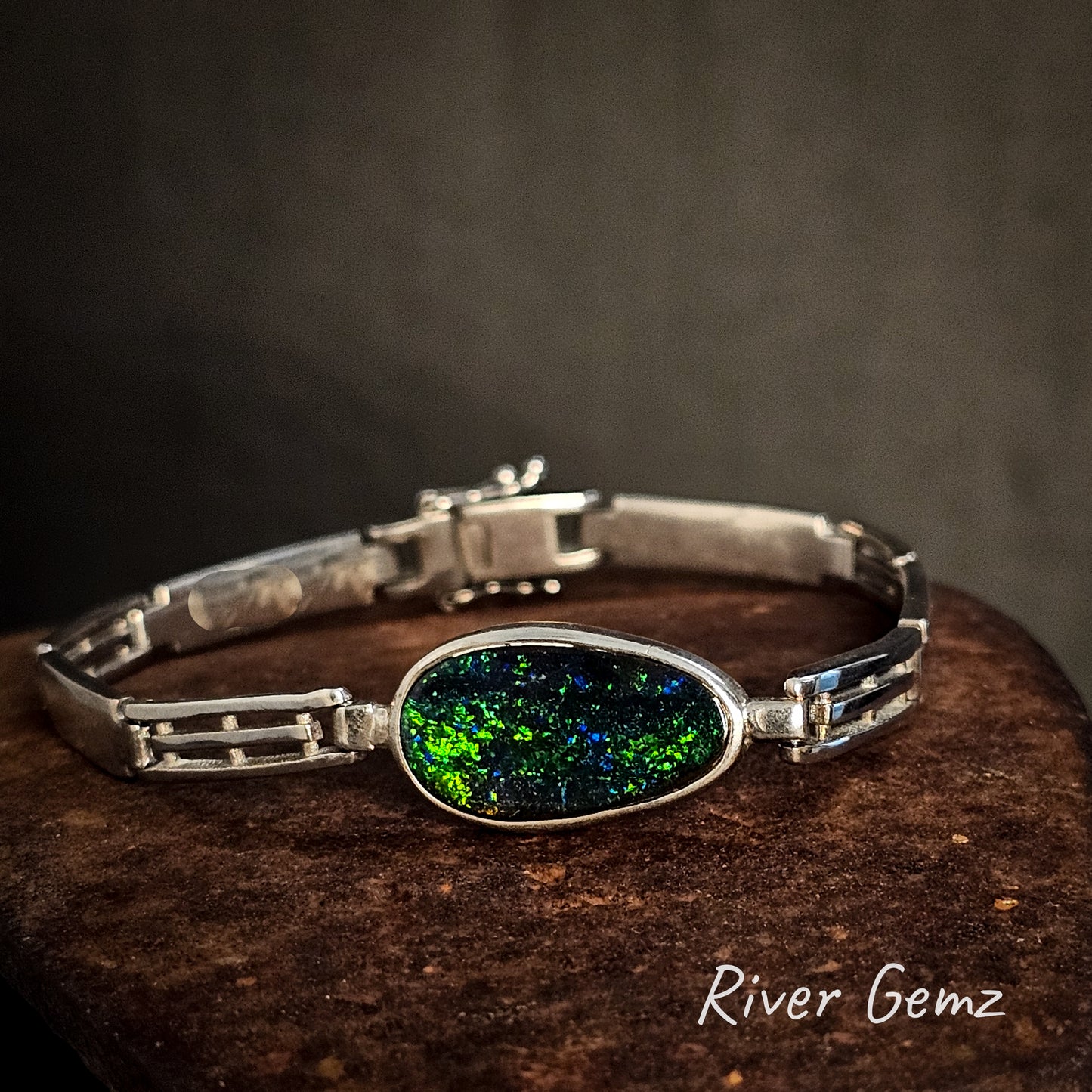 Silver bracelet with a colorful boulder opal on a dark surface, branded 'River Gemz'.
