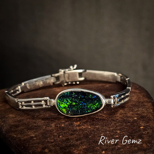 Silver bracelet with a colorful boulder opal on a dark surface, branded 'River Gemz'.