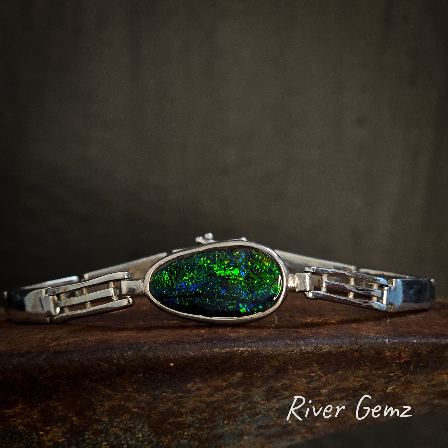 Silver bracelet with a free form cut green and blue opal on a dark background, branded 'River Gemz'.