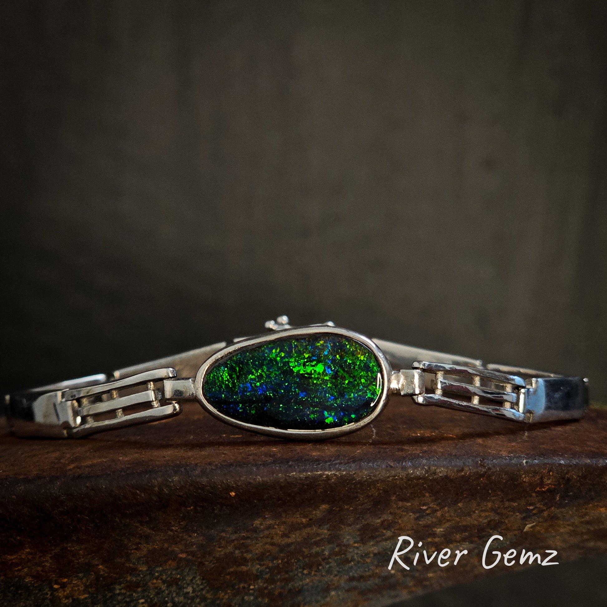 Silver bracelet with a free form cut green and blue opal on a dark background, branded 'River Gemz'.