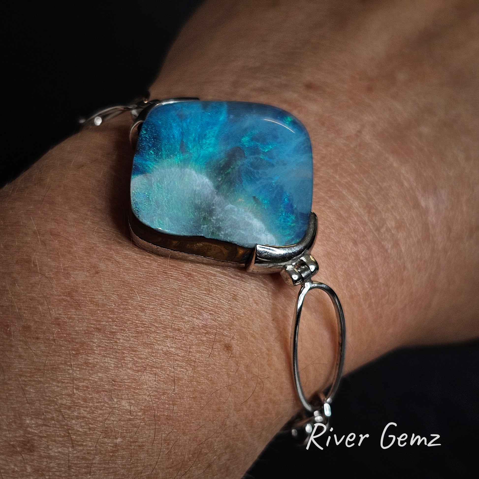 Large turquoise boulder opal in a silver bracelet on a wrist with 'River Gemz' branding.