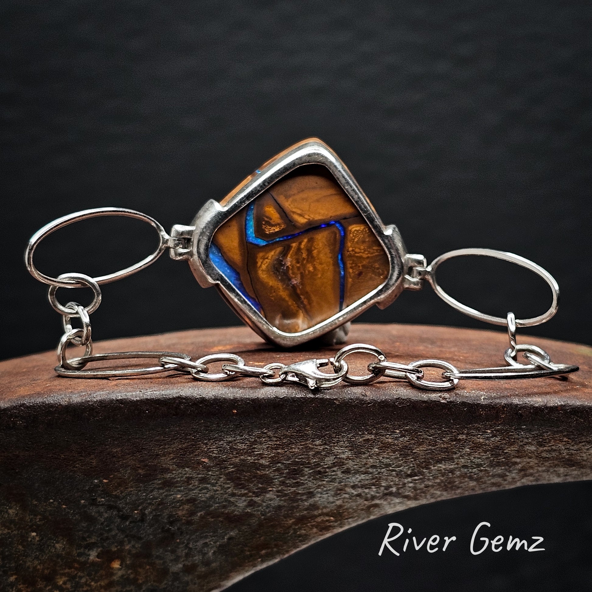 Rear view of silver bracelet showing the ironstone matrix of the boulder opal on a dark background, featuring the brand 'River Gemz'.