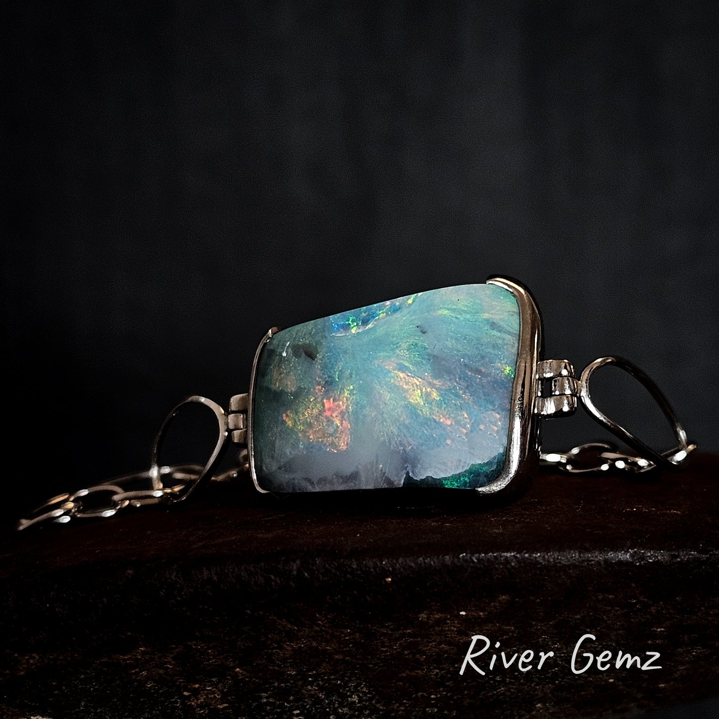 Silver bracelet with a colourful gemstone on a dark background, featuring the brand 'River Gemz'.