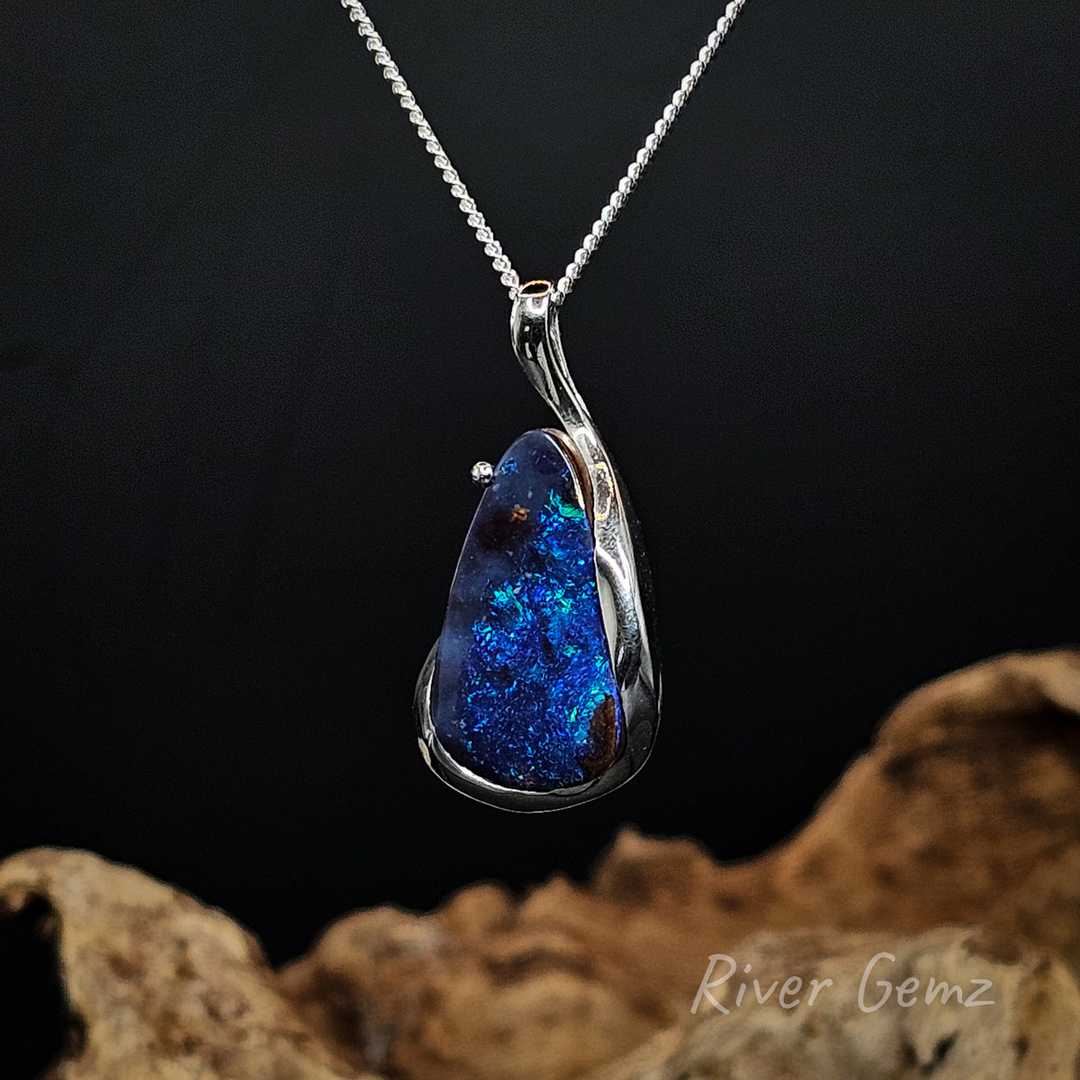 A silver pendant necklace featuring a teardrop-shaped blue opal gemstone with vibrant iridescent flashes, suspended from a delicate silver chain against a dark background with a piece of driftwood at the bottom. The brand name 'River Gemz' is visible in the bottom right corner.
