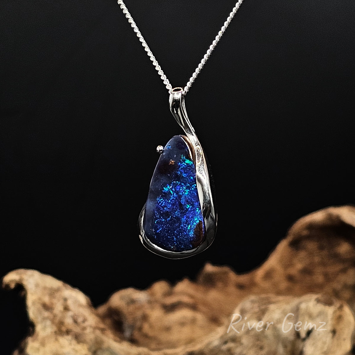 Tear drop shaped blue boulder opal set in silver necklace against a black backround.