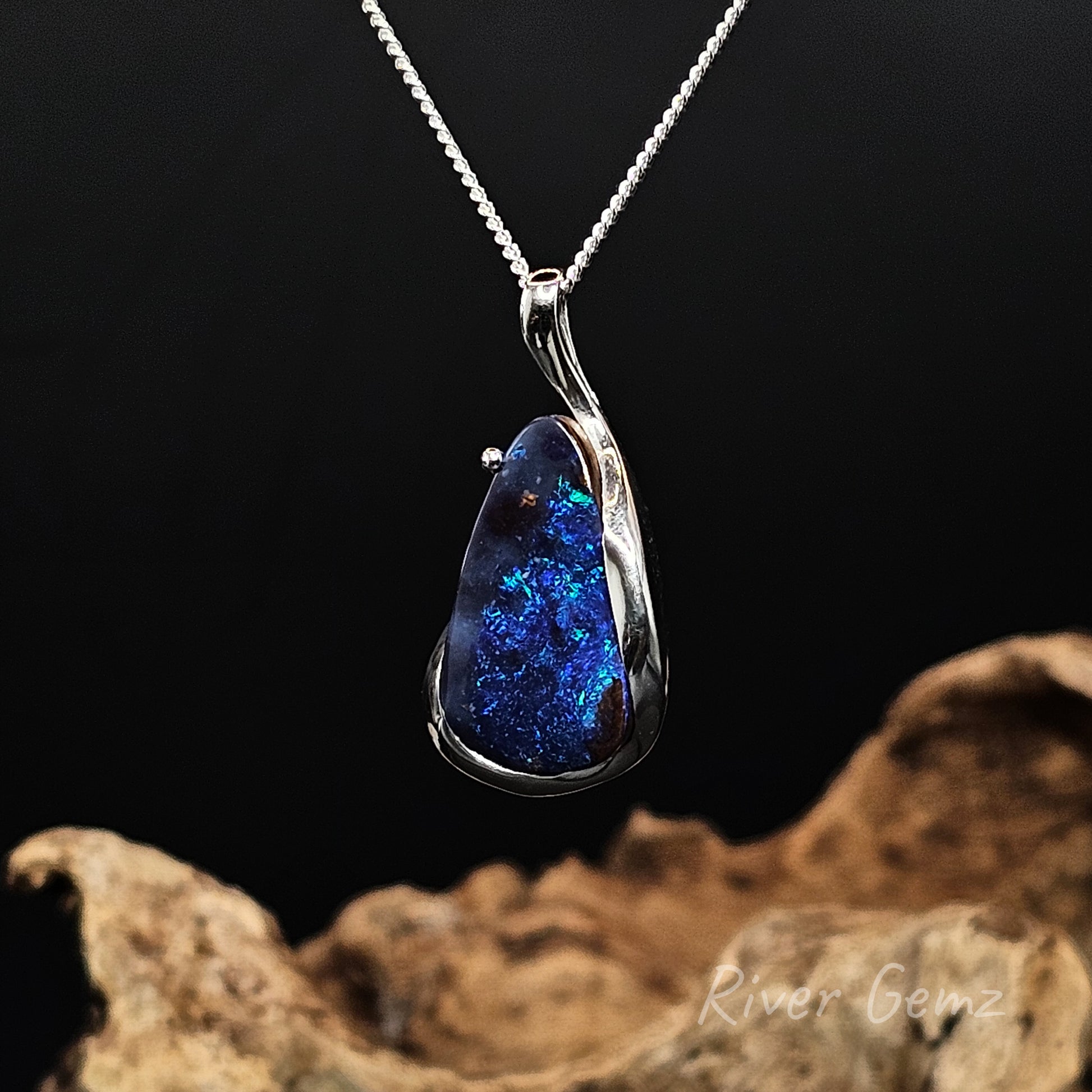 Tear drop shaped blue boulder opal set in silver necklace against a black backround.