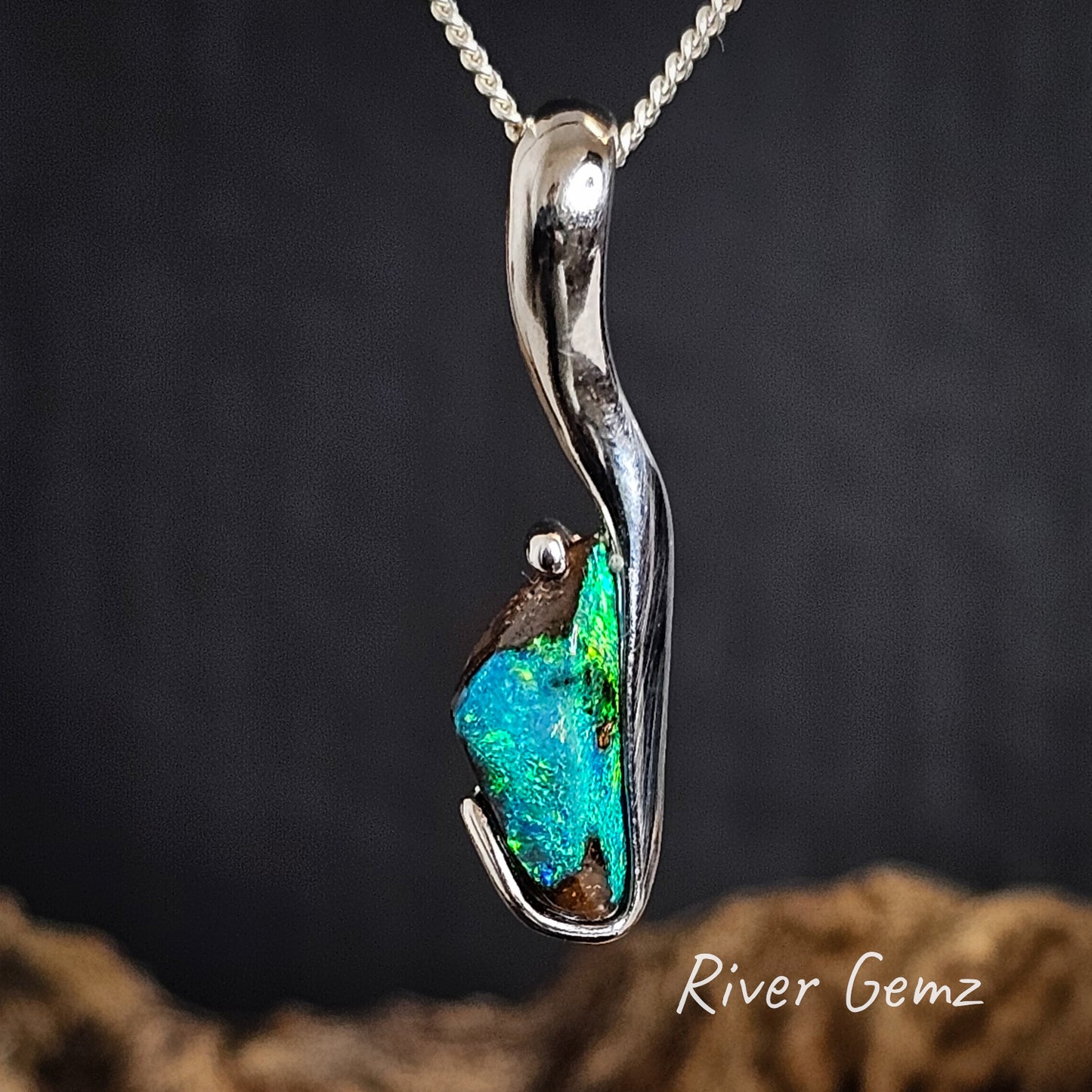 J-shaped silver pendant with a free form cut opal on a dark background, branded 'River Gemz'.