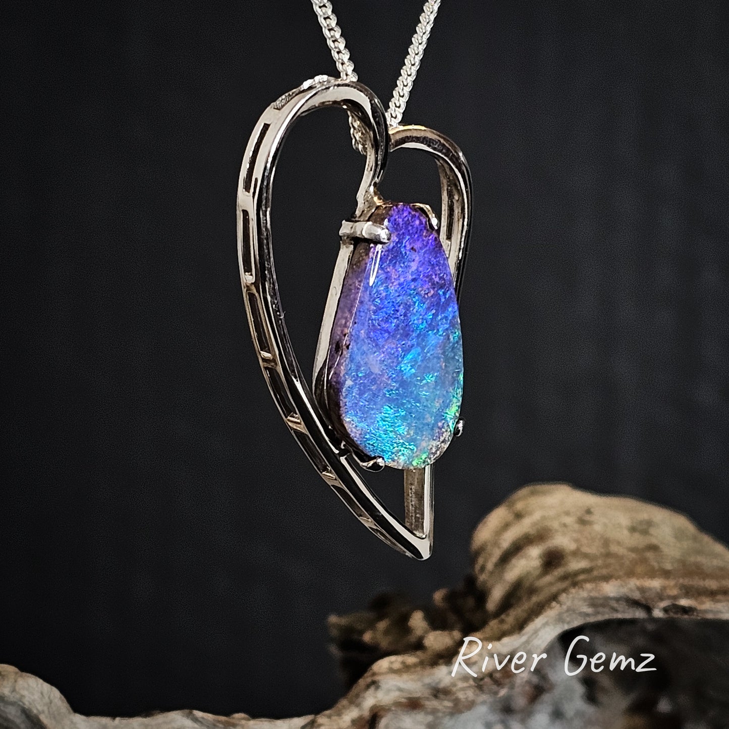 Heart-shaped pendant with a blue, purple and green opal on a dark background, featuring 'River Gemz' branding.