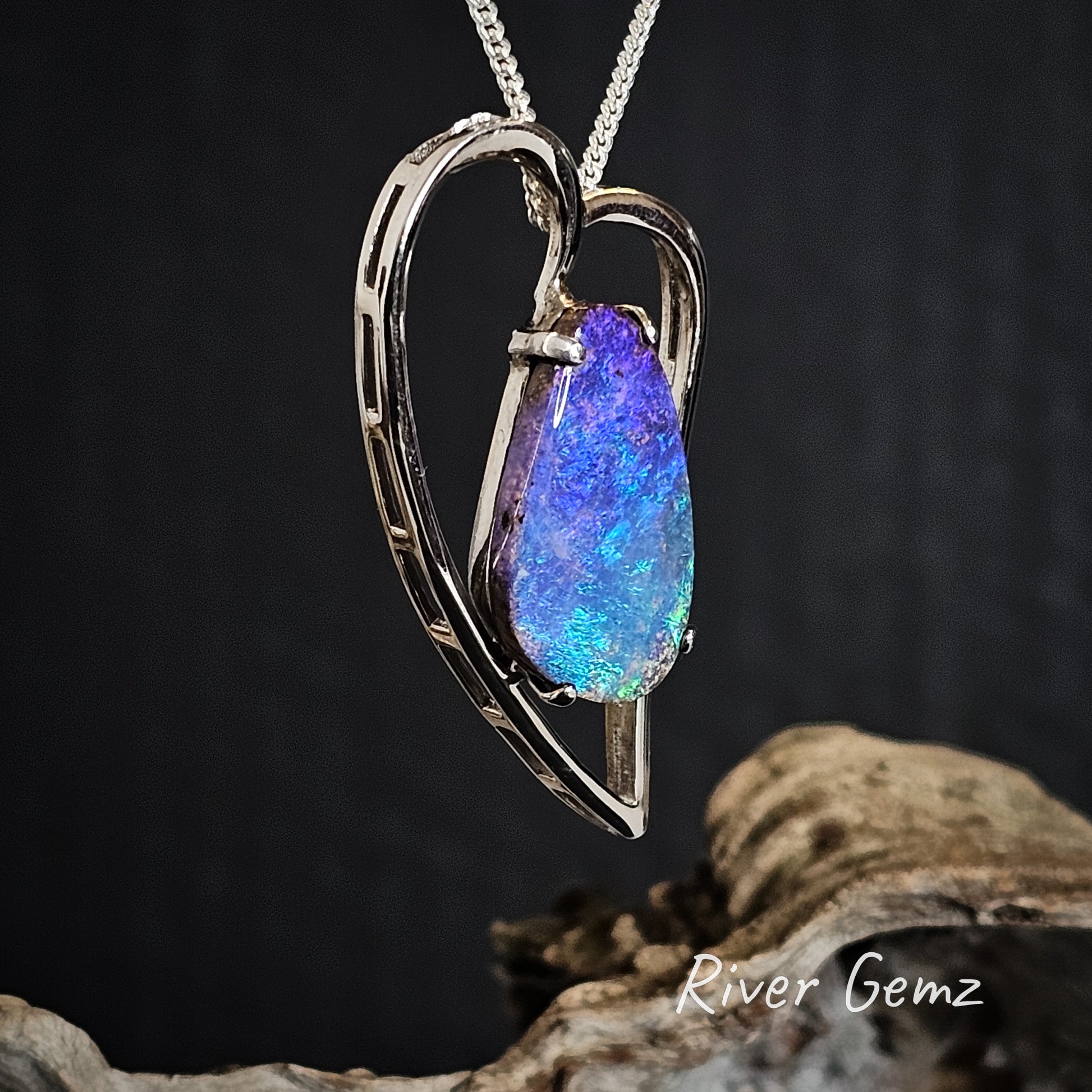Heart-shaped pendant with a blue, purple and green opal on a dark background, featuring 'River Gemz' branding.