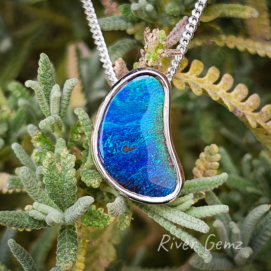 Peanut shaped dark blue and green opal besel set in a silver necklace photographed on lavender bush.