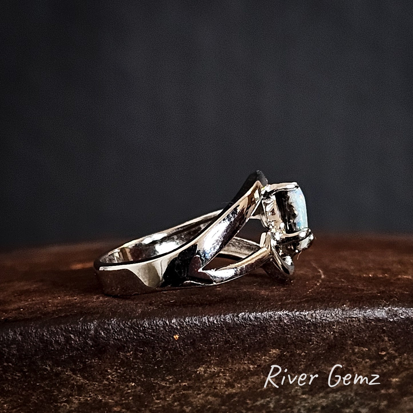 Side view of silver ring with a gemstone on a dark surface, branded 'River Gemz'.