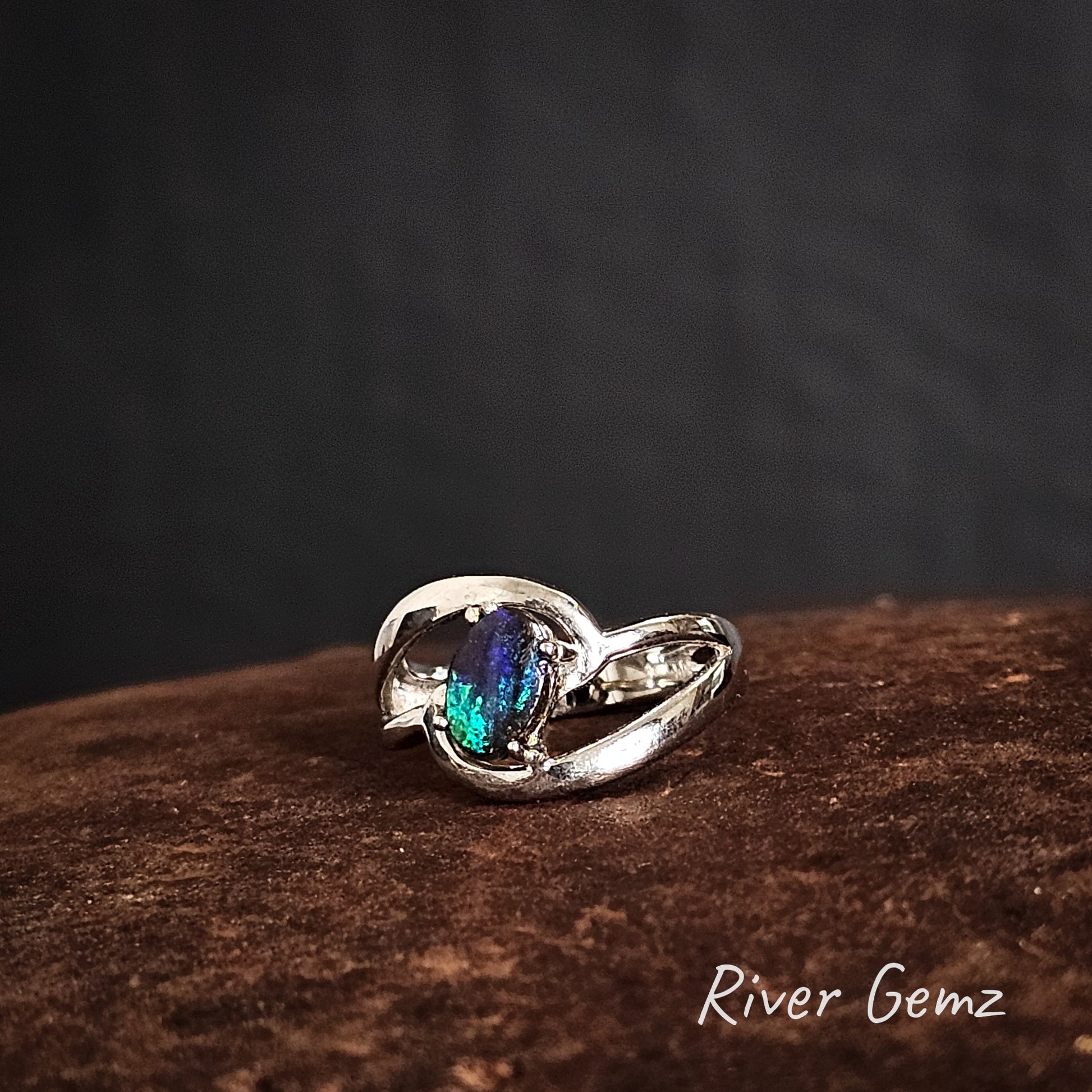 Silver ring with a colorful gemstone on a dark background, featuring the brand 'River Gemz'.