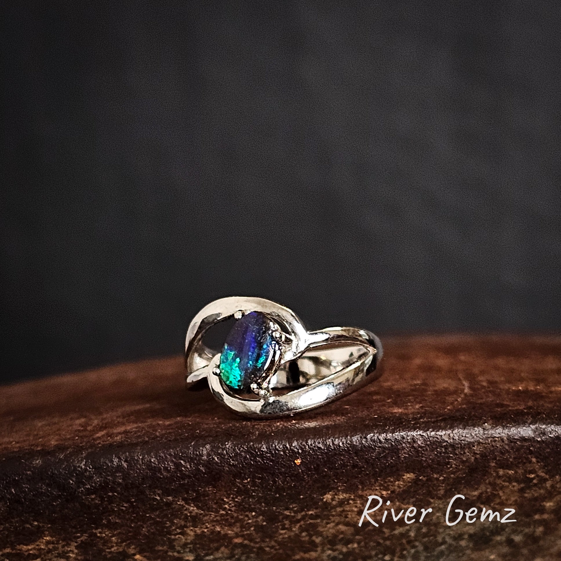 Silver ring with a blue and green boulder opal on a dark cast iron surface, branded 'River Gemz'.