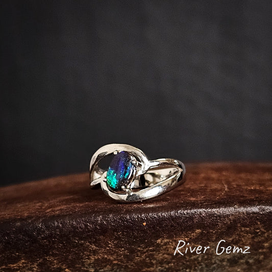 Silver ring with a blue and green boulder opal on a dark cast iron surface, branded 'River Gemz'.