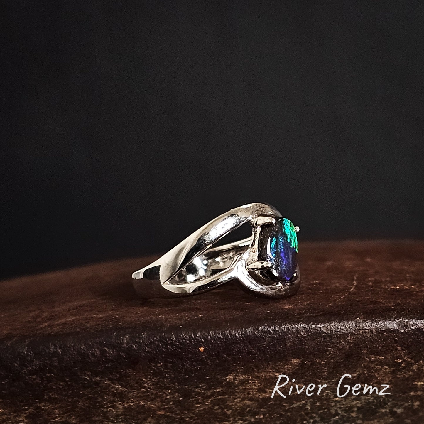 Side view of silver ring with an oval shaped boulder opal on a dark cast iron surface, branded 'River Gemz'.
