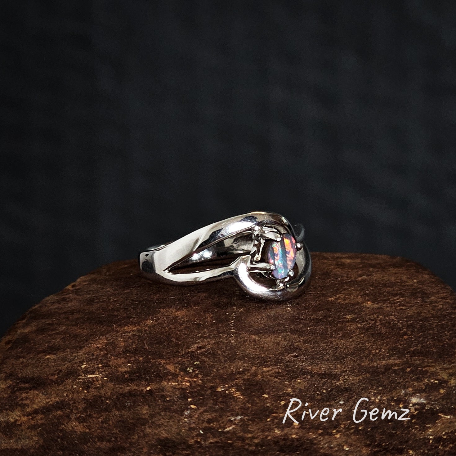 Silver ring with a colorful gemstone on a dark background, branded 'River Gemz'.