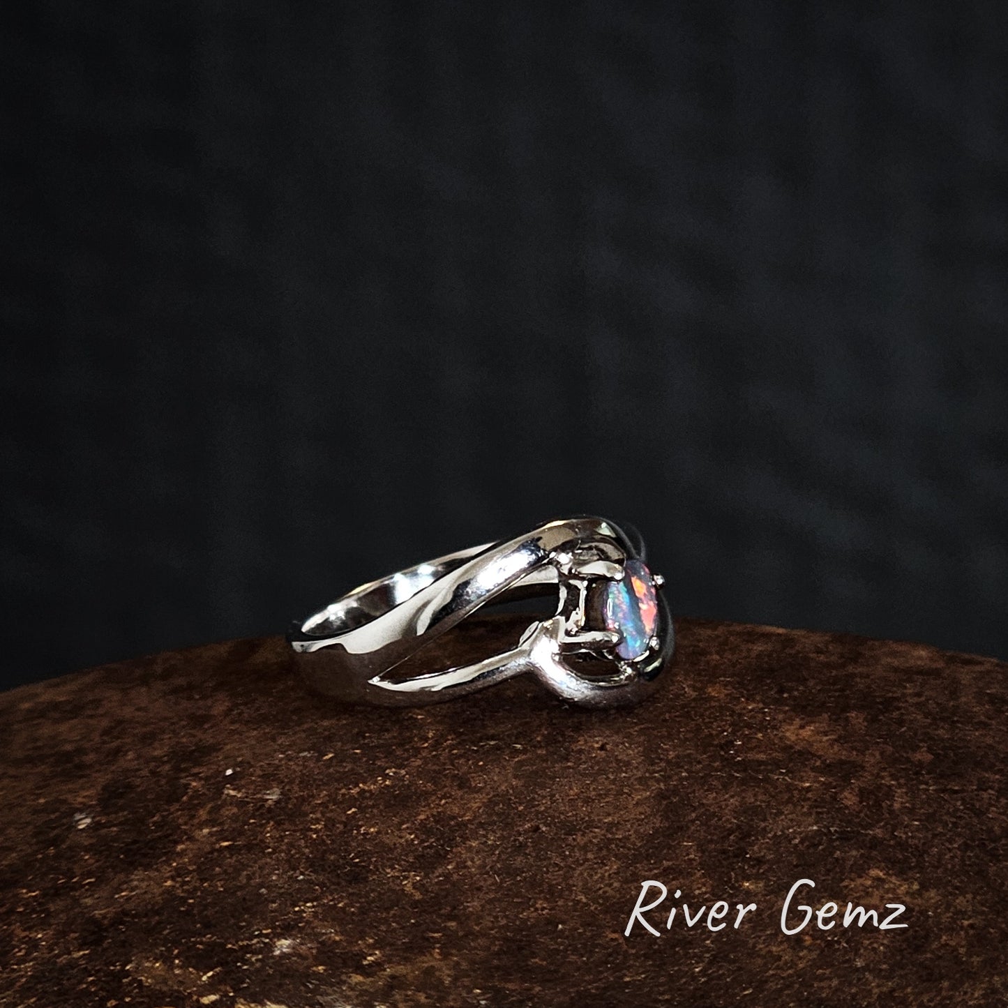 Silver ring with a claw set boulder opal on a dark brown surface, branded 'River Gemz'.