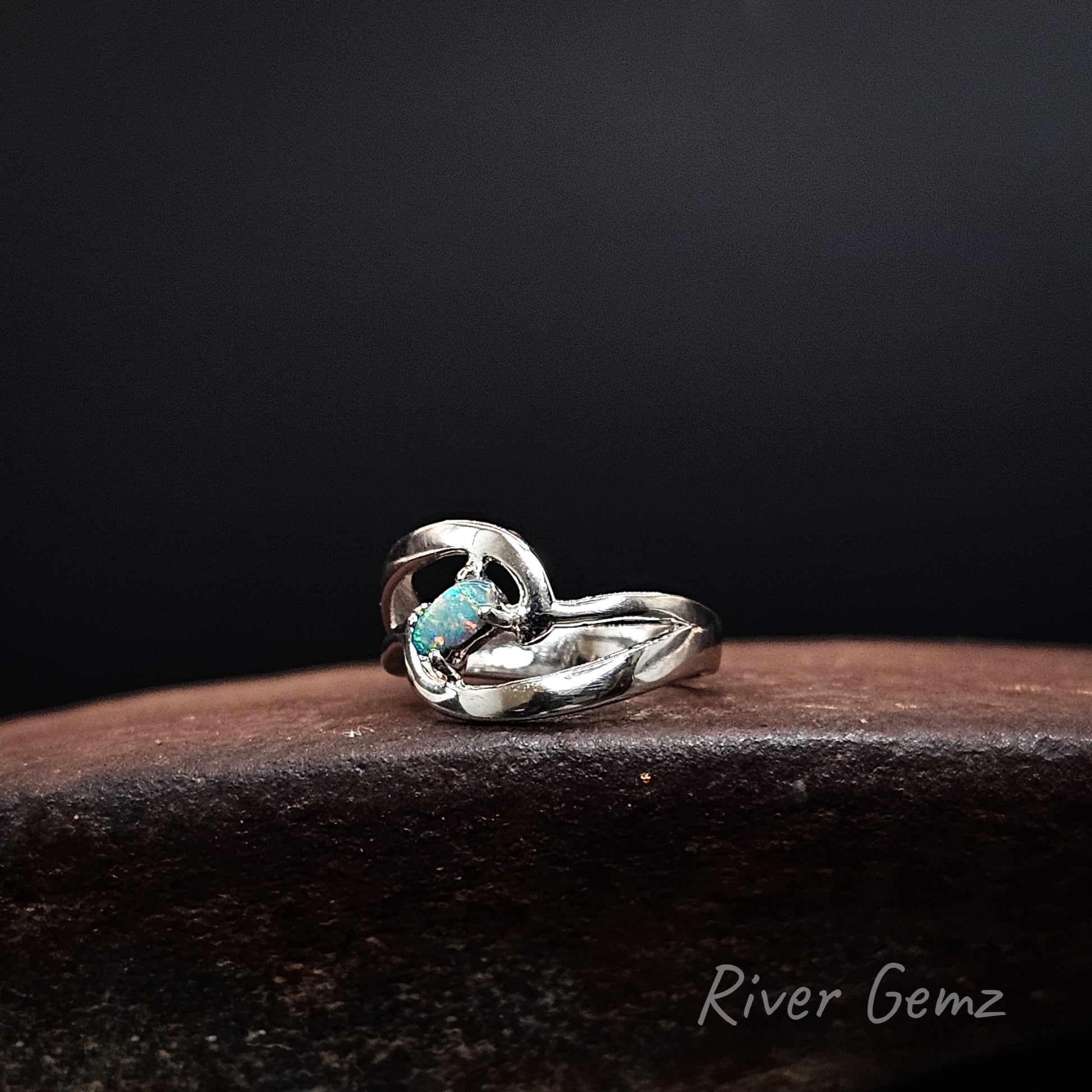 A sterling silver ring with a twisted knot design and a small oval turquoise opal, resting on a dark brown textured surface against a black background. The brand name 'River Gemz' is visible in the bottom right corner.