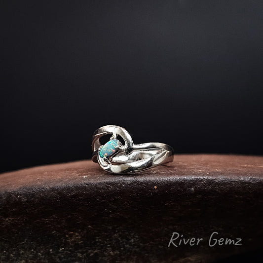 A sterling silver ring with a twisted knot design and a small oval turquoise opal, resting on a dark brown textured surface against a black background. The brand name 'River Gemz' is visible in the bottom right corner.