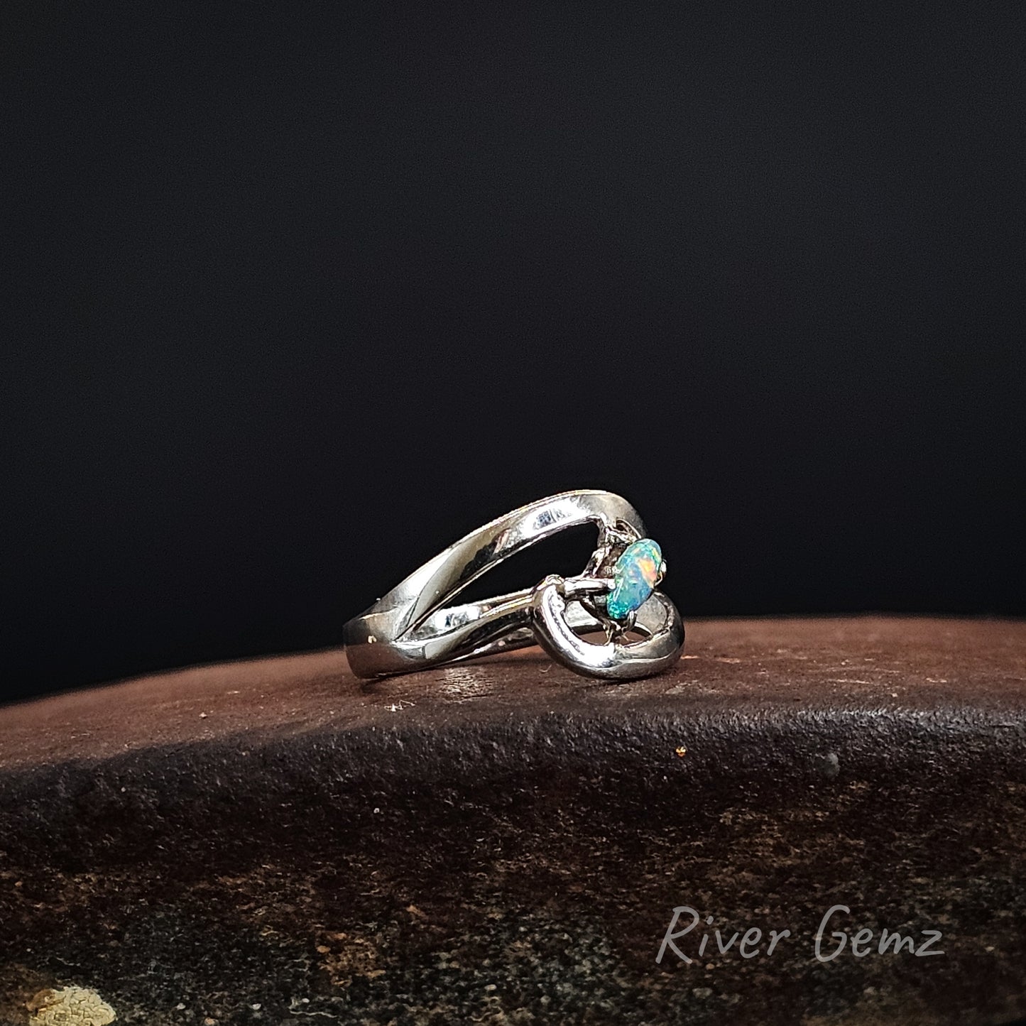 A silver ring with a twisted, openwork band and a small round turquoise-colored gemstone, resting on a dark brown textured surface against a solid black background. The brand name 'River Gemz' is watermarked in the bottom right corner.