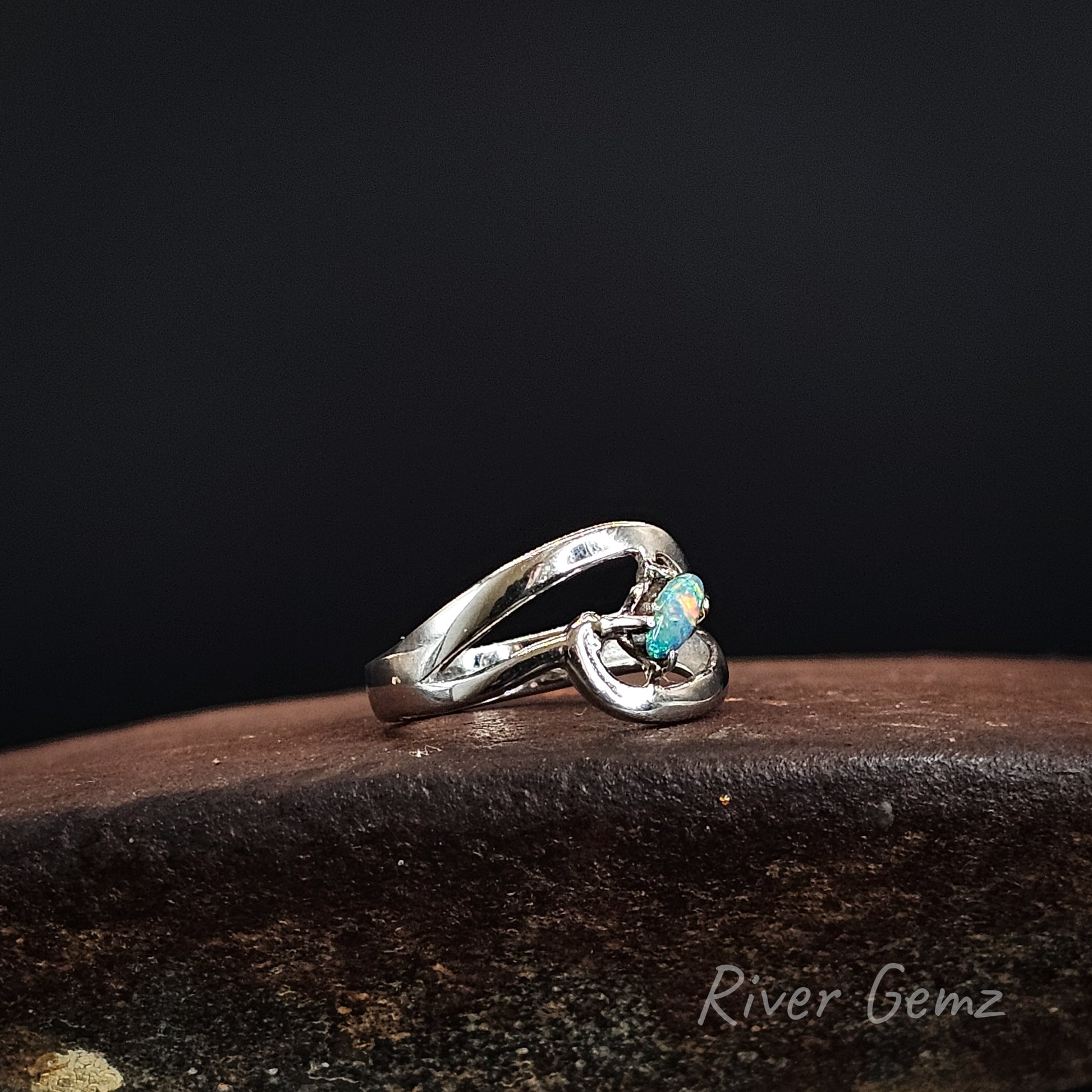 A silver ring with a twisted, openwork band and a small round turquoise-colored gemstone, resting on a dark brown textured surface against a solid black background. The brand name 'River Gemz' is watermarked in the bottom right corner.