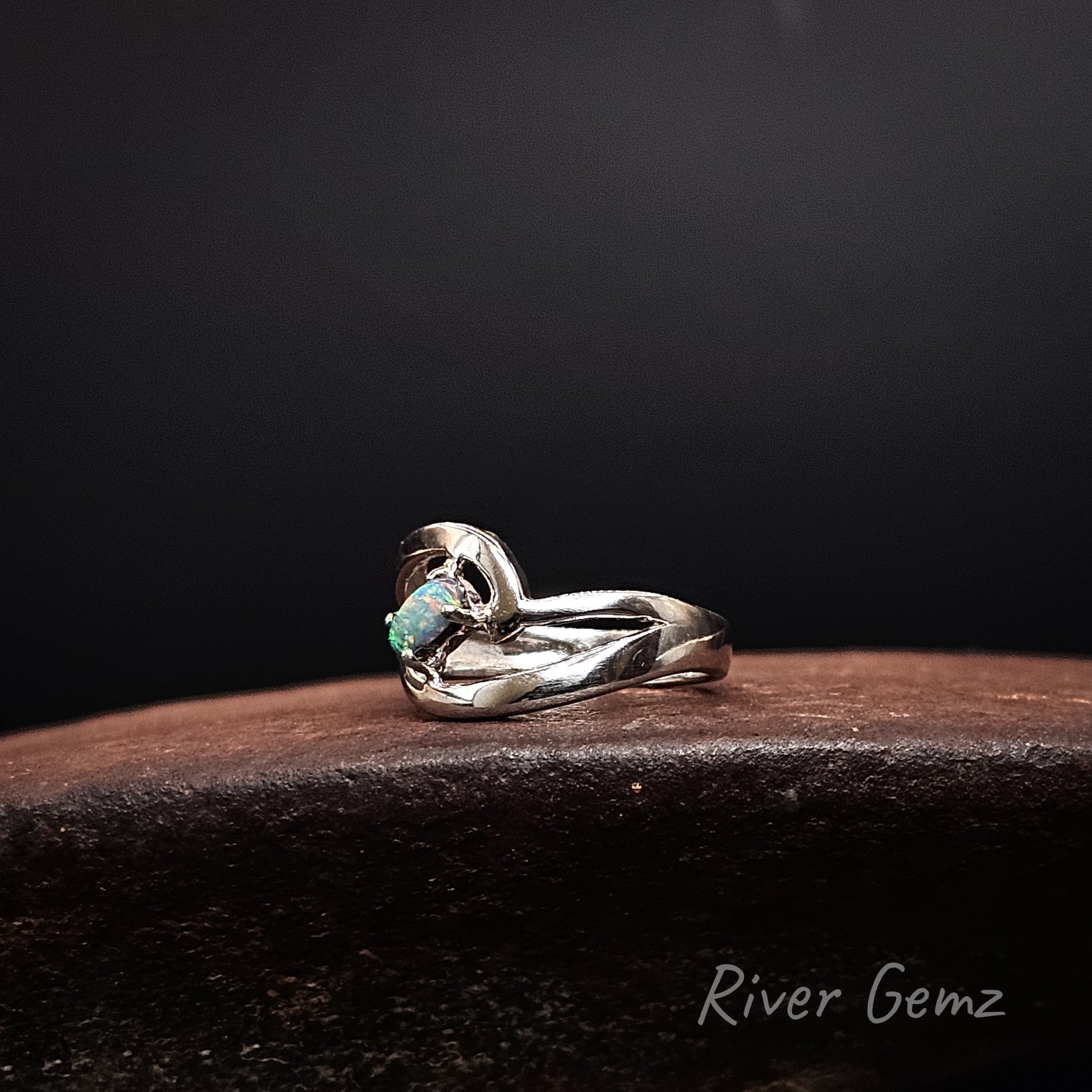 A silver ring with a curved, textured band and a small green opal gemstone, displayed on a dark brown wooden surface against a black background. The brand name 'River Gemz' is visible in the bottom right corner.