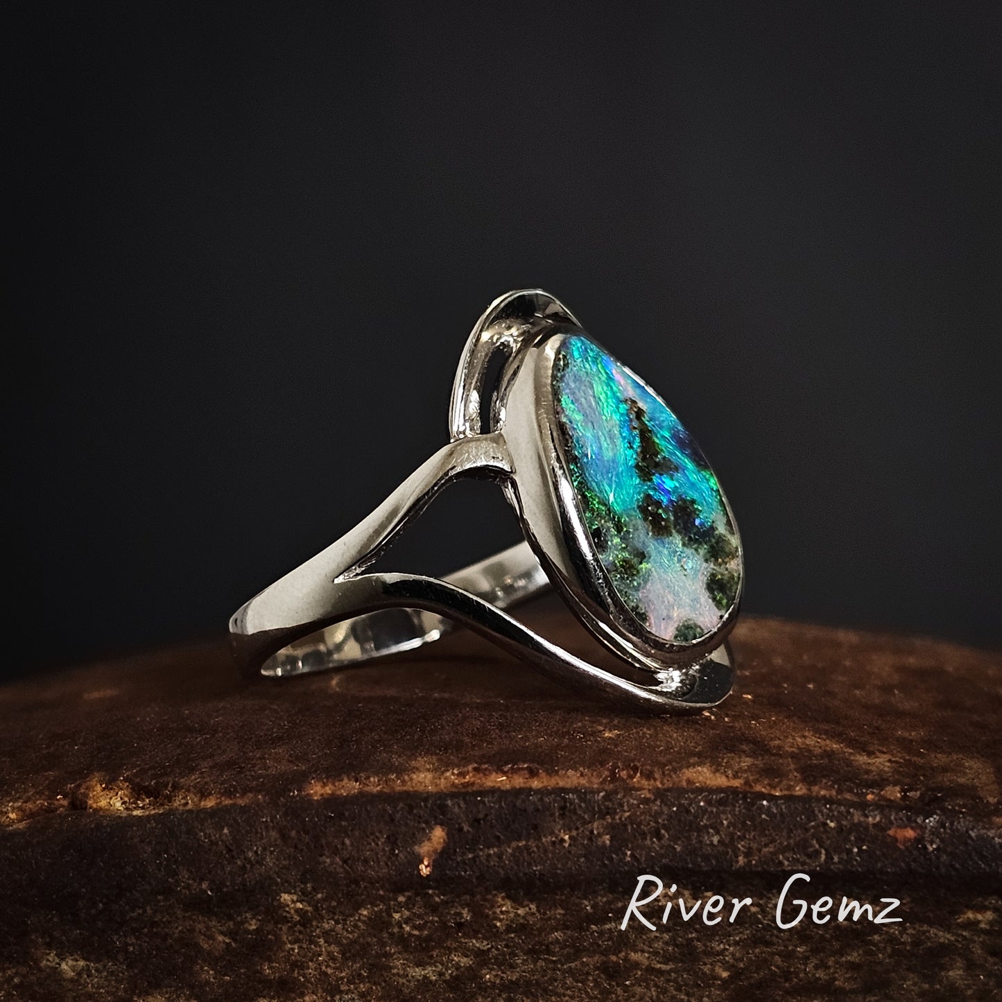 Silver ring with a colourful night sky like boulder opal on a dark background, branded 'River Gemz'.