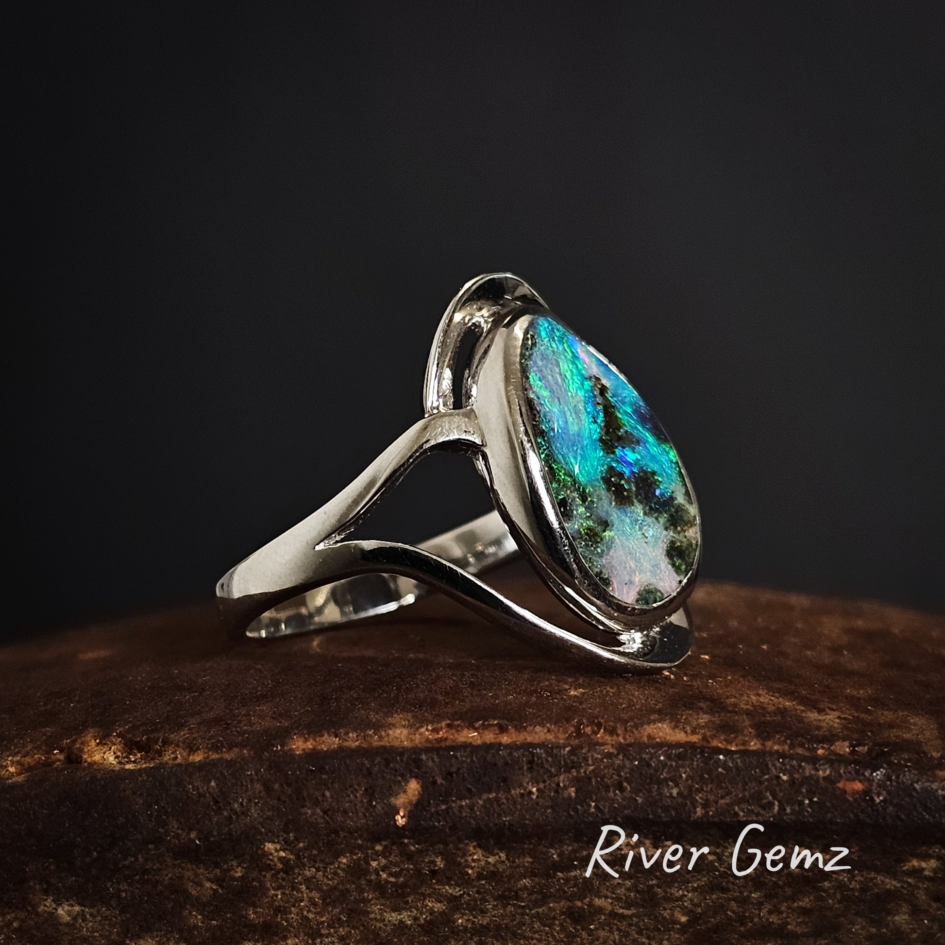 Silver ring with a colourful night sky like boulder opal on a dark background, branded 'River Gemz'.