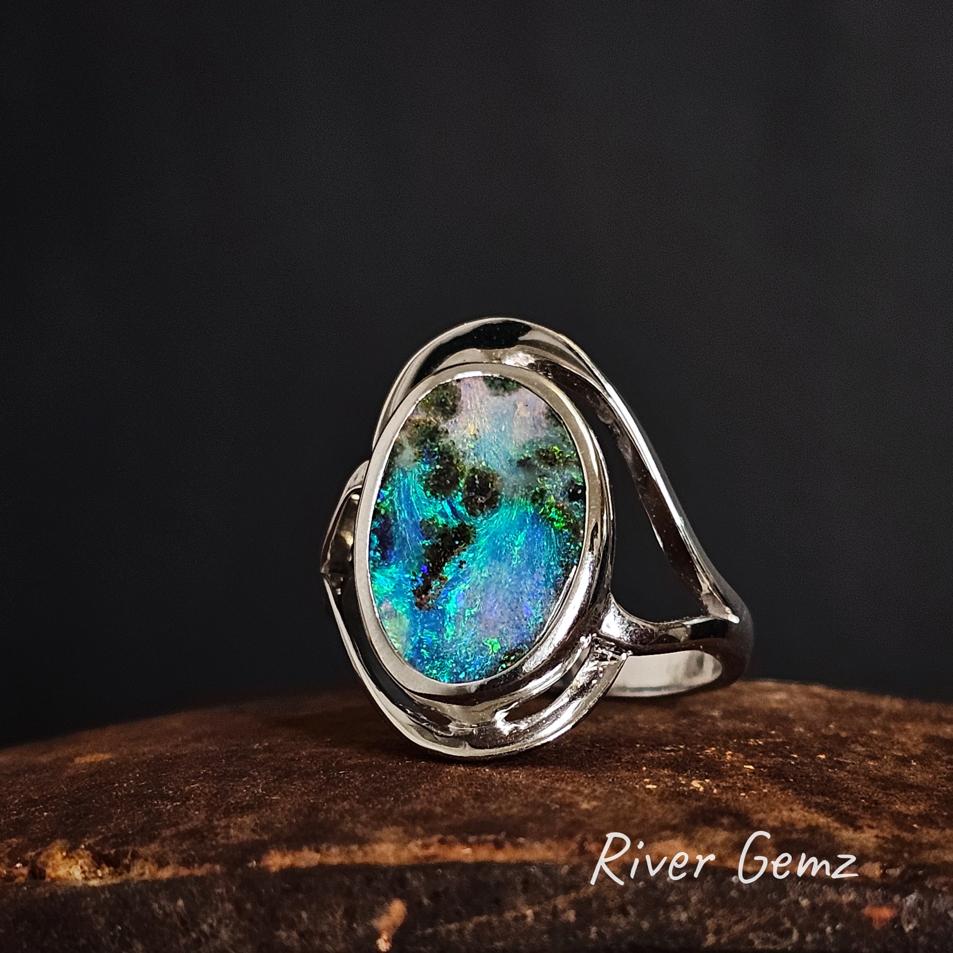 Silver ring with a colorful gemstone on a dark background, branded 'River Gemz'.