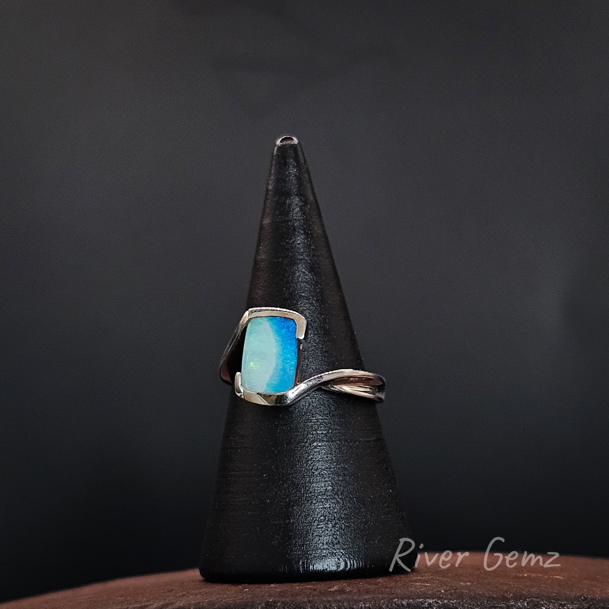 A silver ring with a rectangular light blue cabochon gemstone, displayed on a black cone stand against a dark background. The brand name 'River Gemz' is visible as a watermark in the bottom right corner.