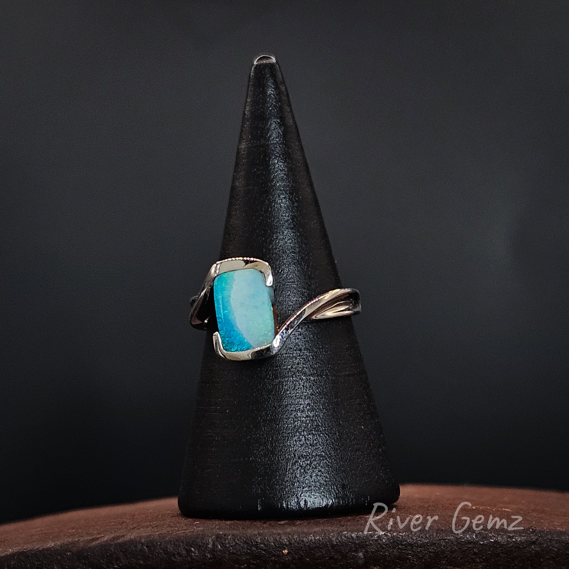 A silver ring with a rectangular turquoise-blue gemstone, displayed on a black cone stand against a dark background. The brand name 'River Gemz' is visible as a watermark in the bottom right corner.