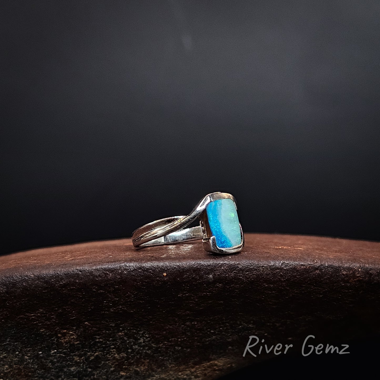 A sterling silver ring with a rectangular turquoise-blue gemstone, resting on a dark wooden surface against a dark gray background. The brand name 'River Gemz' is visible as a watermark in the bottom right corner.