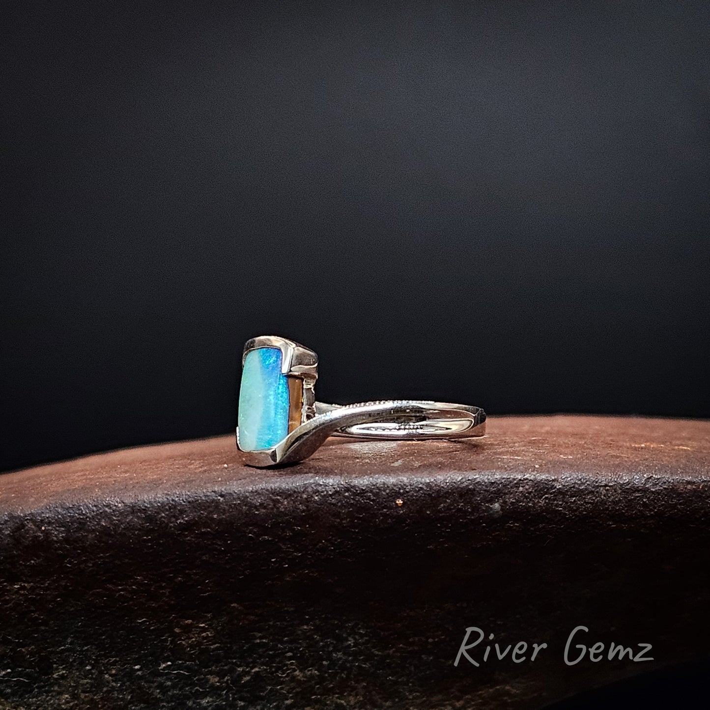 A silver ring featuring a rectangular blue opal gemstone, resting on a dark textured stone surface against a dark gray background. The brand name 'River Gemz' is visible in the bottom right corner.