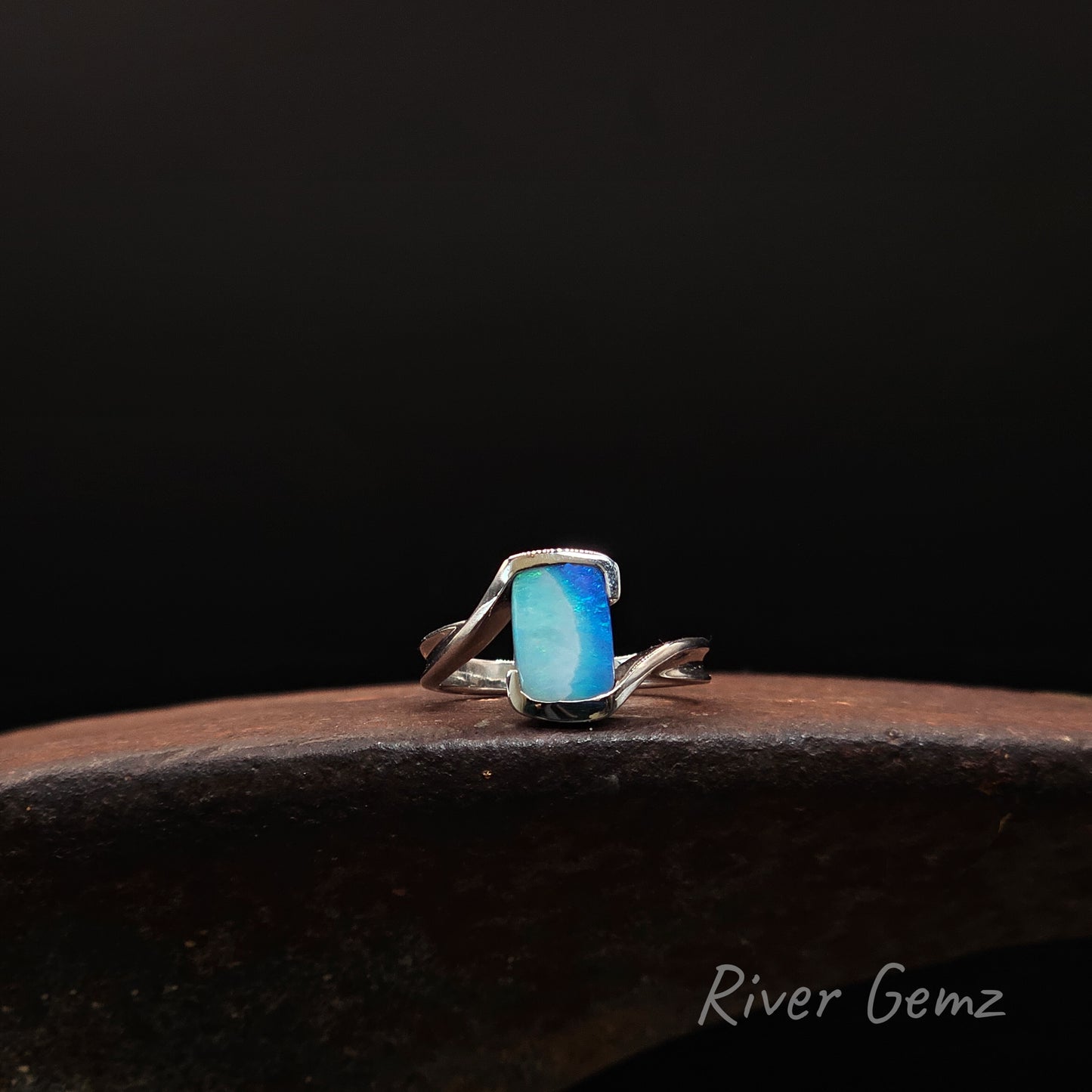 A silver ring with a rectangular blue-green stone, displayed on a dark wooden surface against a black background. The text 'River Gemz' is visible in the bottom right corner.