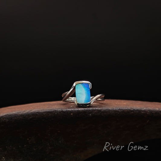 A silver ring with a rectangular blue-green stone, displayed on a dark wooden surface against a black background. The text 'River Gemz' is visible in the bottom right corner.