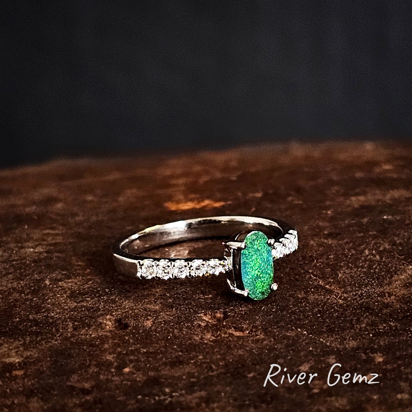 Silver ring with a bright green boulder opal and 5 white topaz stones on each shoulder on a dark brown surface, branded 'River Gemz'.