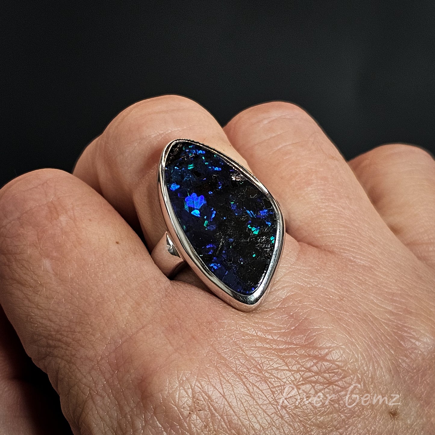 Silver ring with dark opal worn on the middle finger of a hand.
