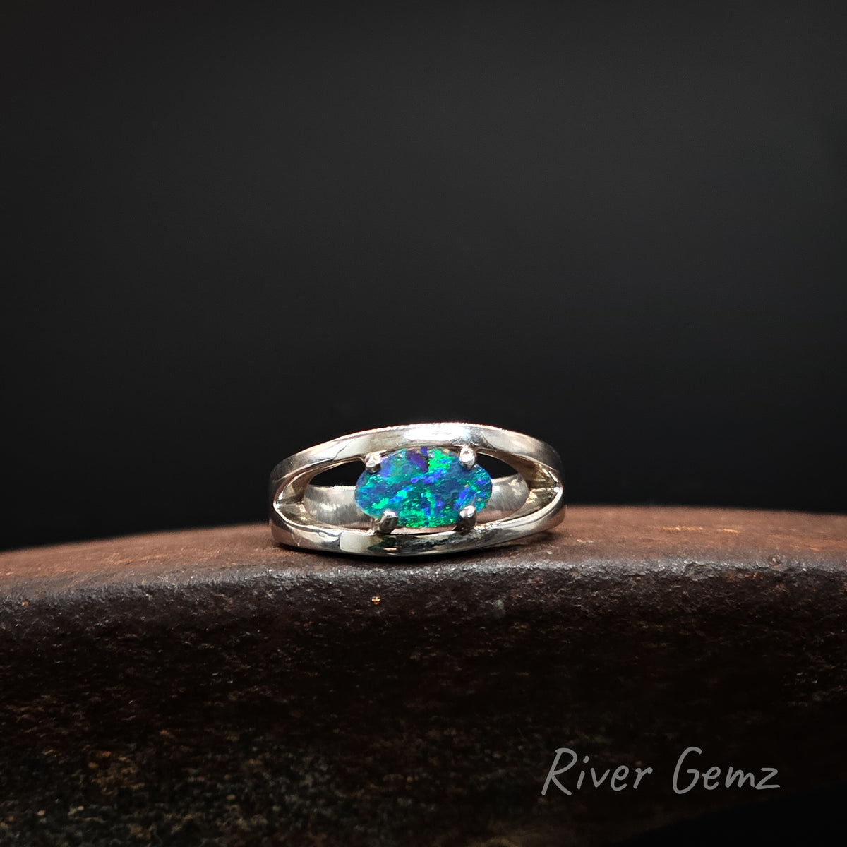 A sterling silver ring with a vibrant blue-green opal gemstone, resting on a dark textured surface against a black background. The brand name 'River Gemz' is visible in the bottom right corner.