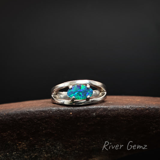 A sterling silver ring with a vibrant blue-green opal gemstone, resting on a dark textured surface against a black background. The brand name 'River Gemz' is visible in the bottom right corner.