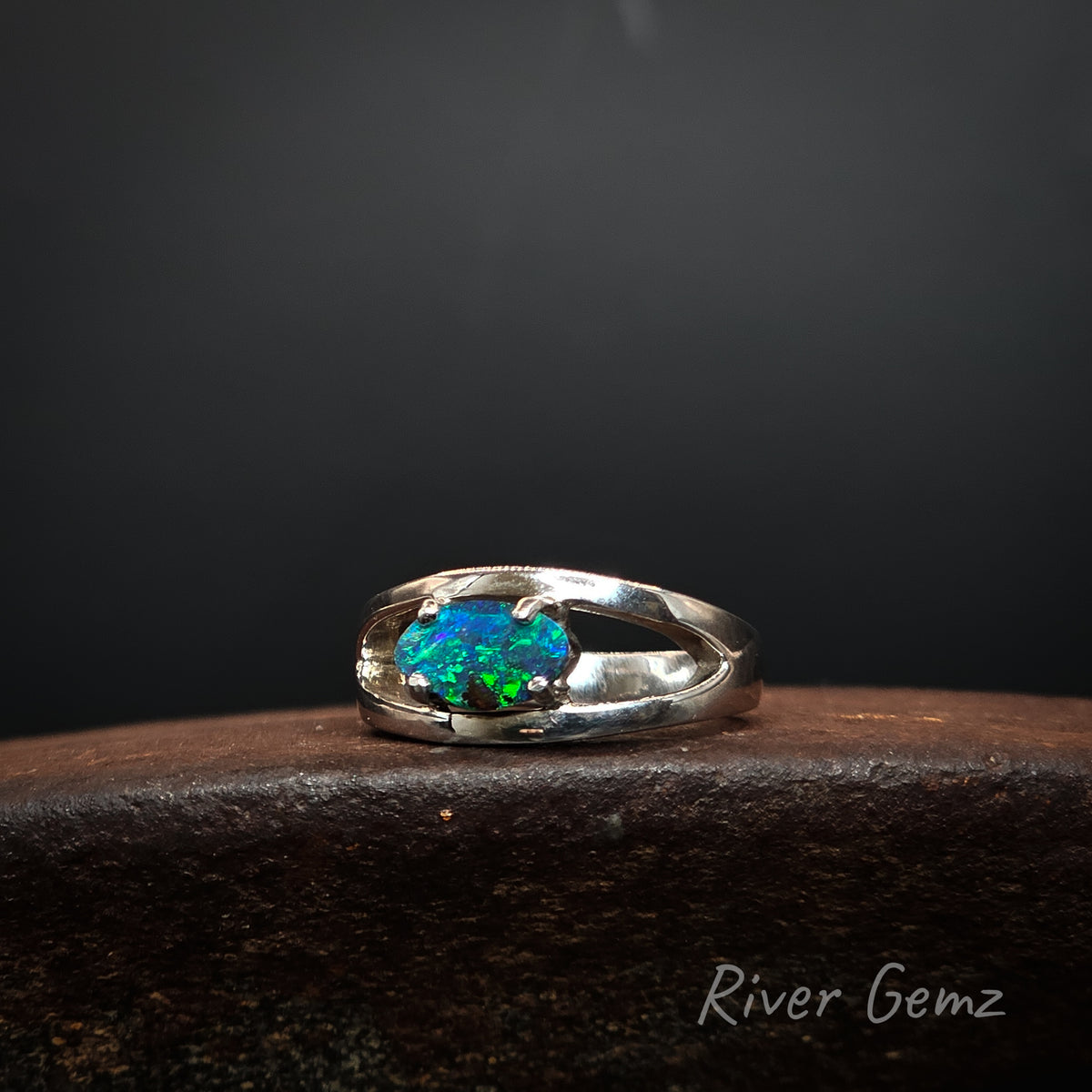 A sterling silver ring featuring a vibrant, multicolored oval opal gemstone, displayed on a dark textured surface against a dark gray background. The brand name 'River Gemz' is visible in the bottom right corner.